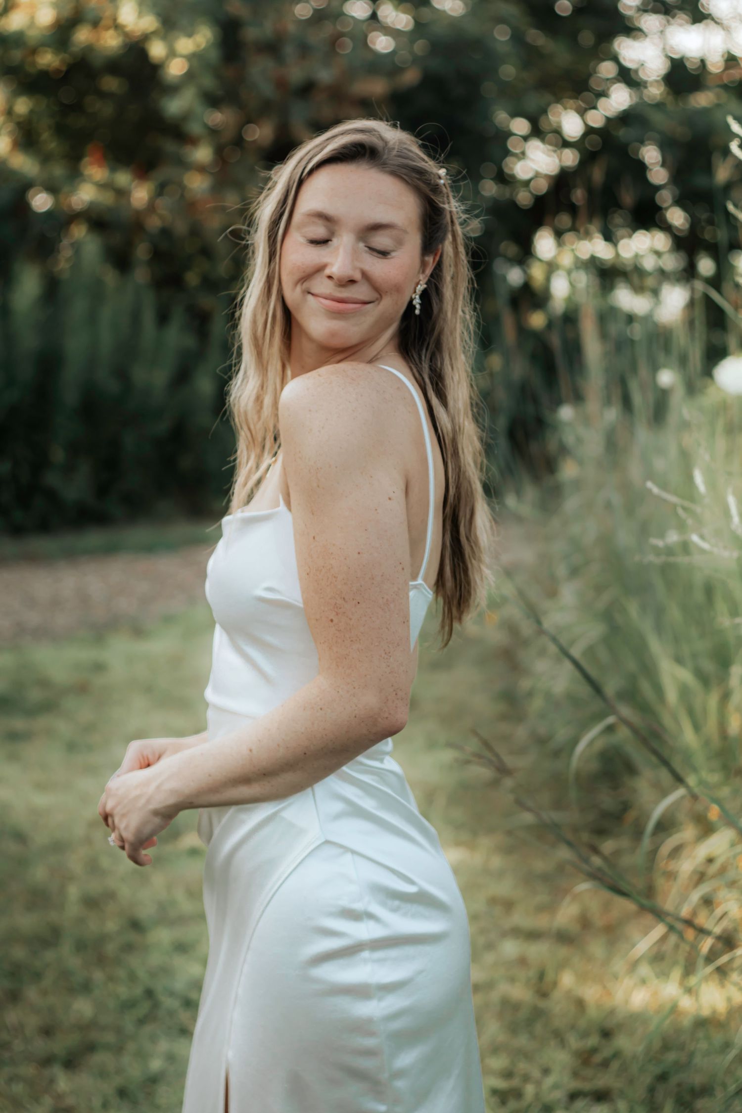 A person wearing a white dress poses outdoors in a natural garden setting with soft sunlight filtering through trees.
