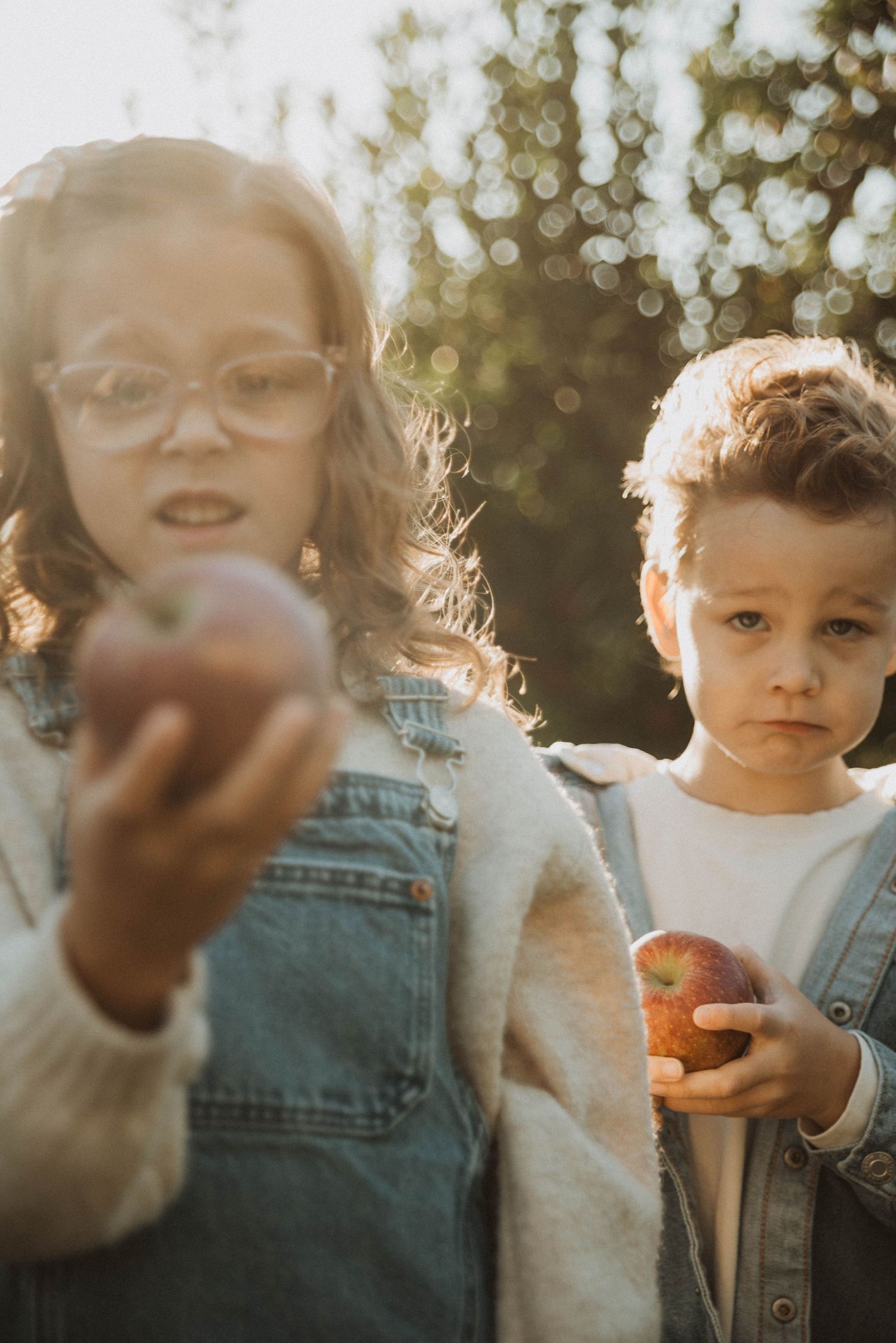 Two children holding red apples outdoors in golden sunlight, girl with glasses extending apple toward camera.