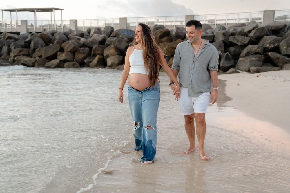 Pregnant couple holding hands walking barefoot on a sandy beach near rocky jetty during golden hour.
