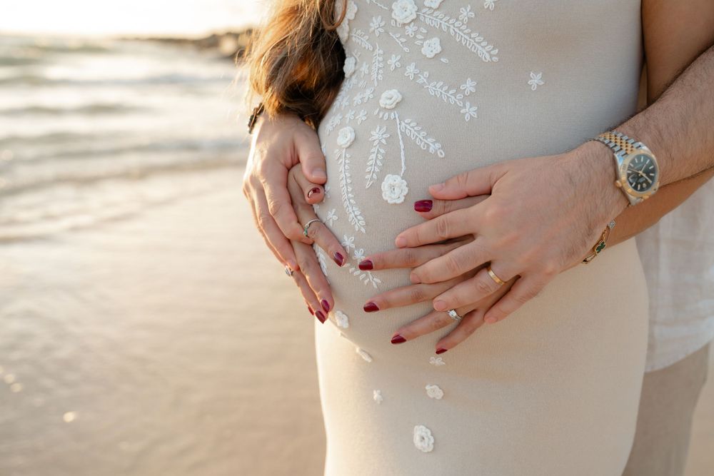 Pregnant woman in white dress cradling baby bump on beach with partner's hands, wearing gold watch and red nails.