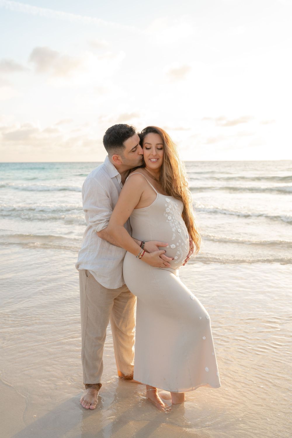 Couple sharing a tender moment on a beach at sunset, man kissing pregnant woman in a beige dress.