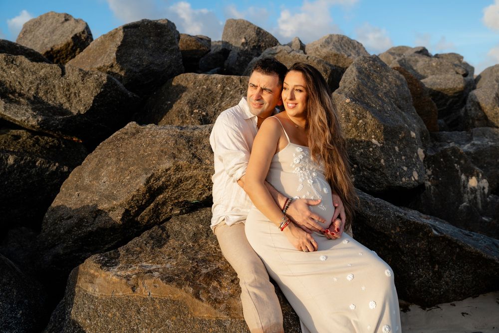 Smiling pregnant couple posing together against large rocks on a sunny beach, woman in white dress.