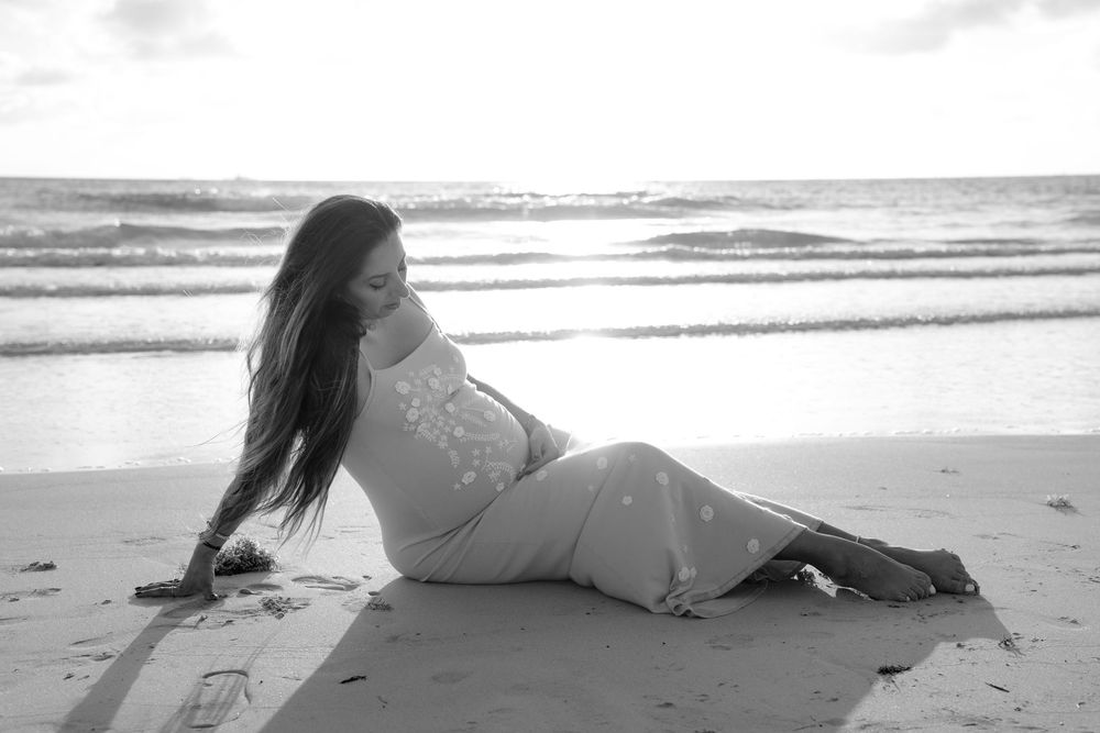 Black and white photo of a woman in a flowing dress sitting gracefully on a sandy beach with ocean waves behind her.