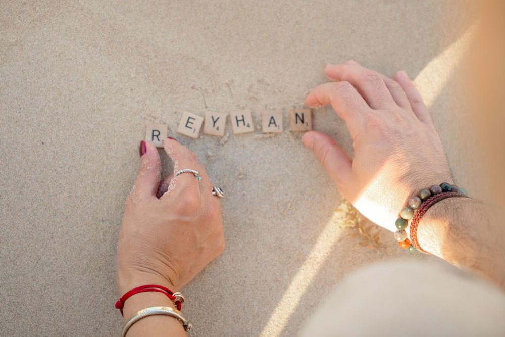 Two hands arranging Scrabble tiles spelling REYHAN on a sandy beach surface in warm sunlight.