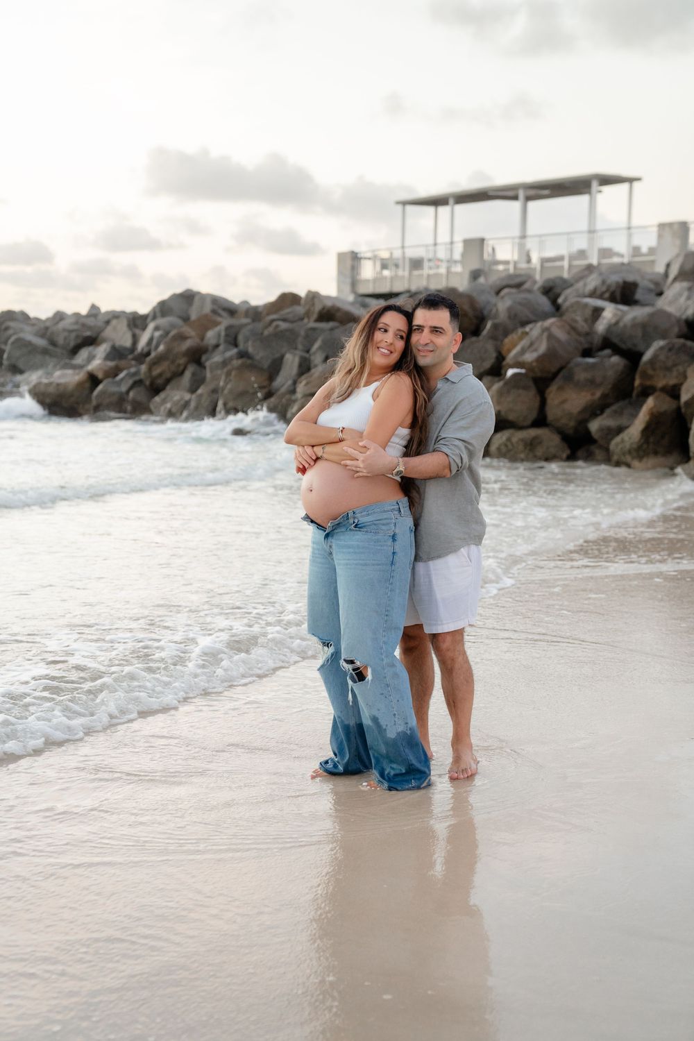 Pregnant couple embracing on a sandy beach at sunset, waves and rocky jetty in the background.