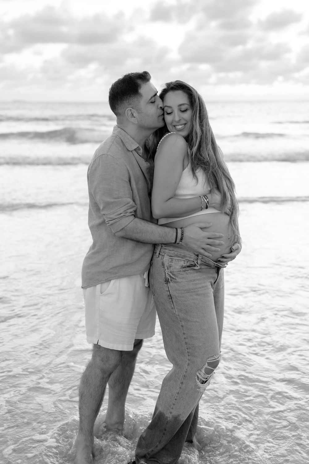 Black and white photo of a couple embracing on a beach, woman visibly pregnant, waves in the background.