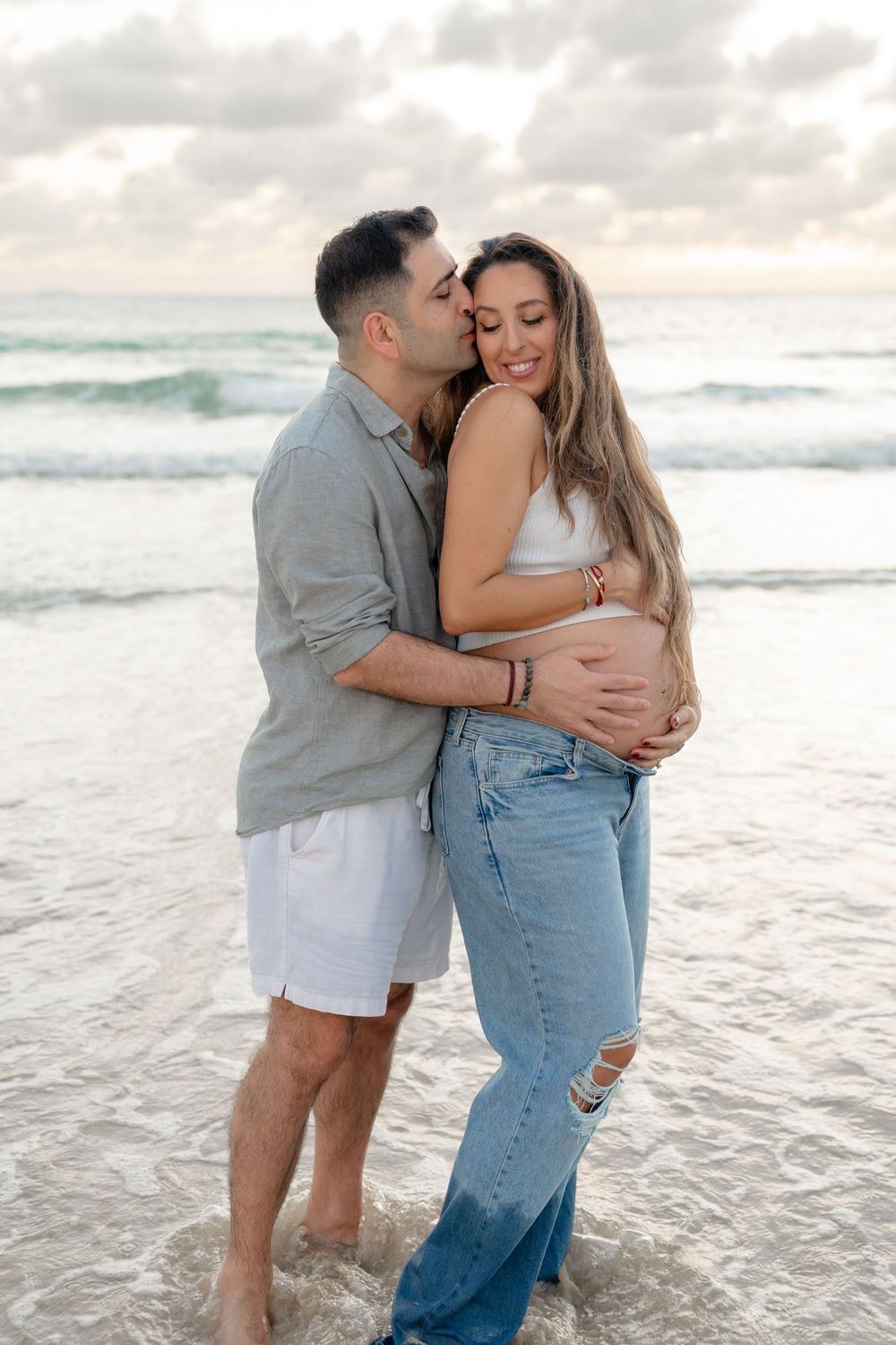 Couple embracing on beach at sunset, pregnant woman smiling as partner kisses her cheek near ocean waves.