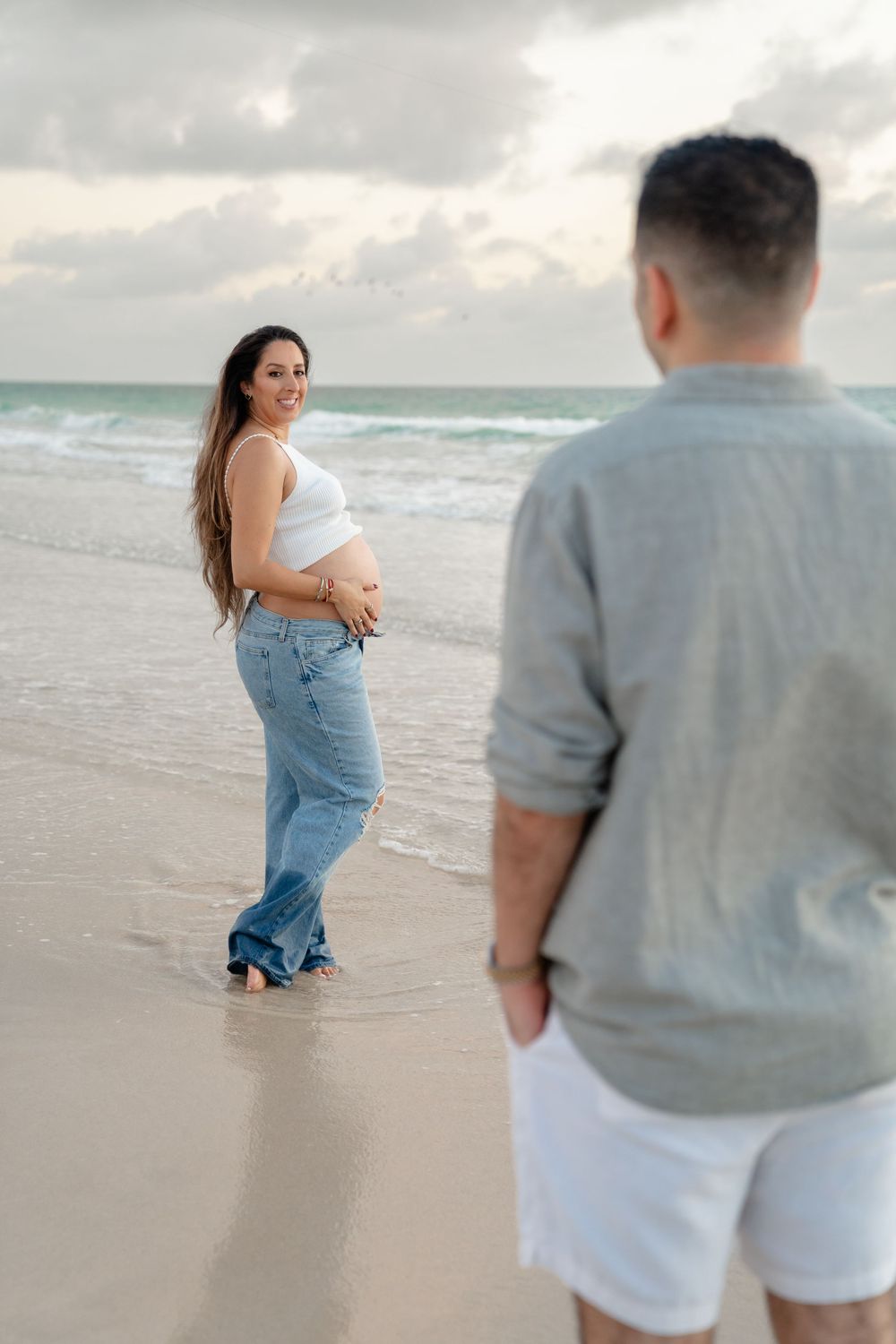 Pregnant woman in white crop top and jeans smiles on beach while man watches from foreground near ocean waves.