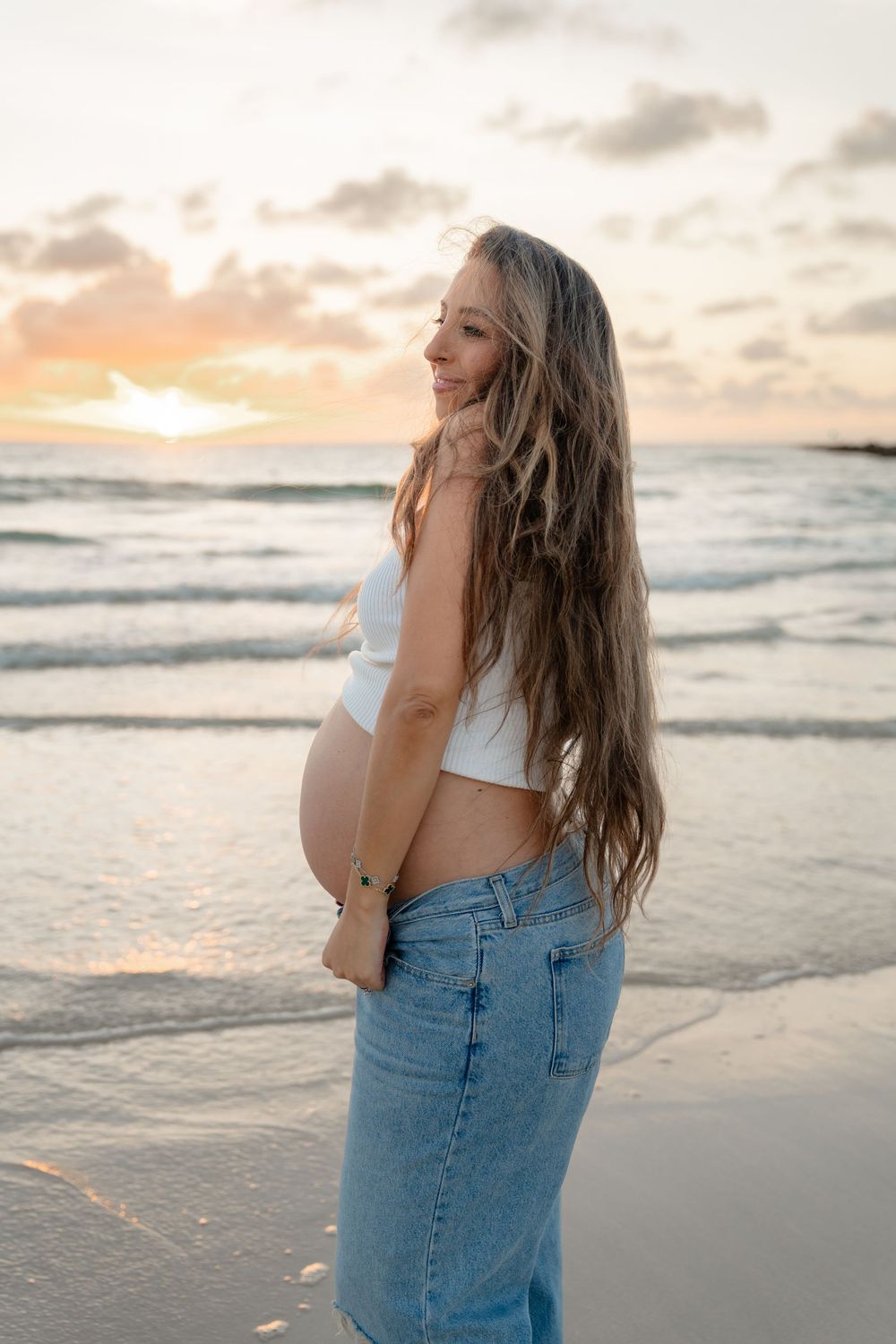 Smiling pregnant woman in white crop top and jeans standing on beach at sunset with long flowing hair.