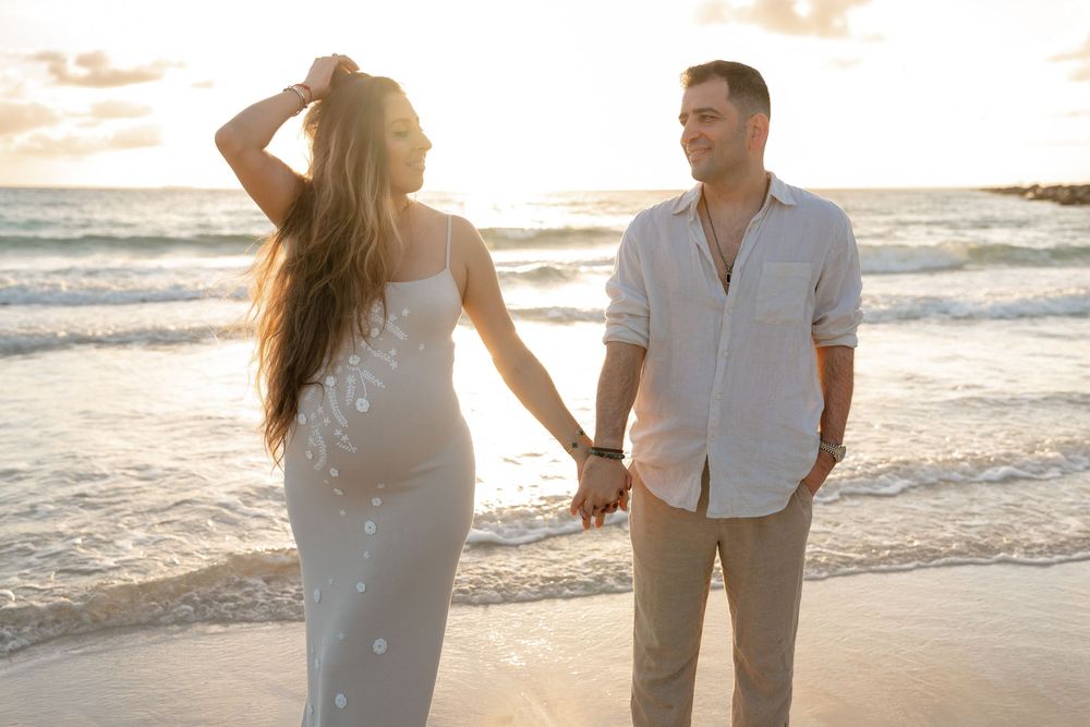 Couple holding hands on a tropical beach at sunset, woman in elegant white dress, man in casual linen shirt.