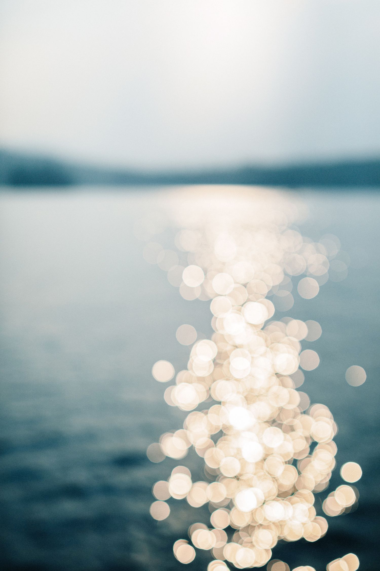 Bokeh light reflections shimmer on blue lake with soft sunlight and distant tree-lined horizon. Southern Maine family vacation photographer.