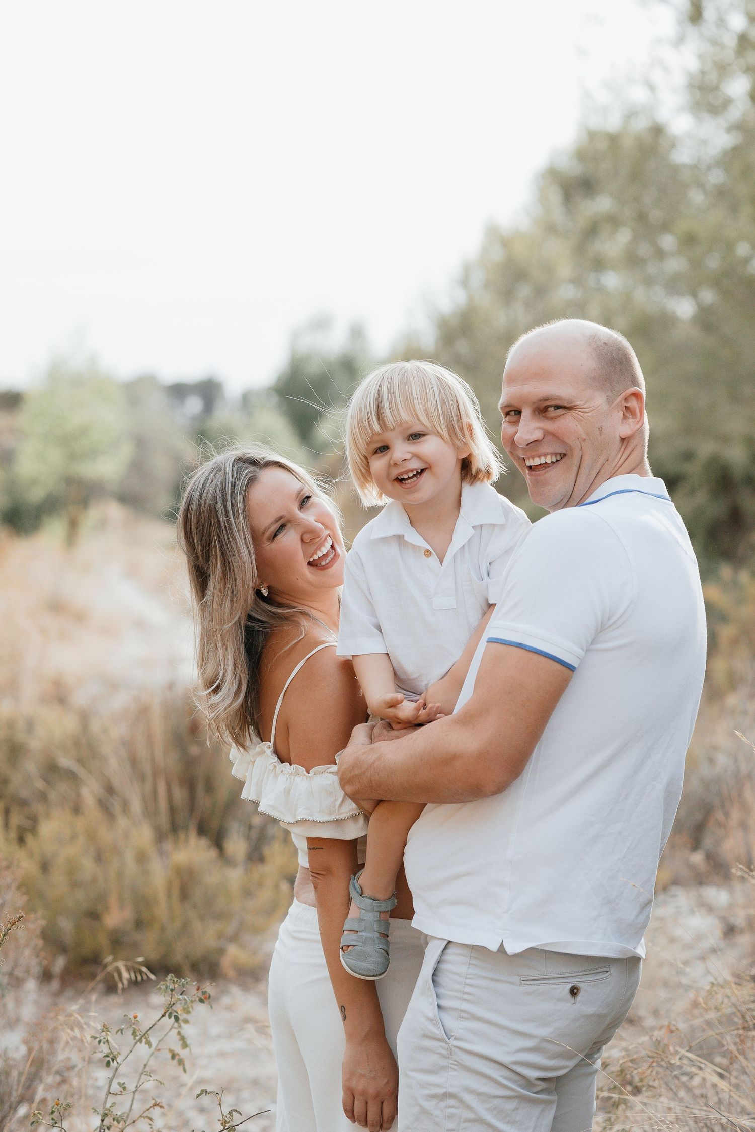 Happy family with laughing toddler son outdoors in sunny field, parents smiling in white summer outfits.