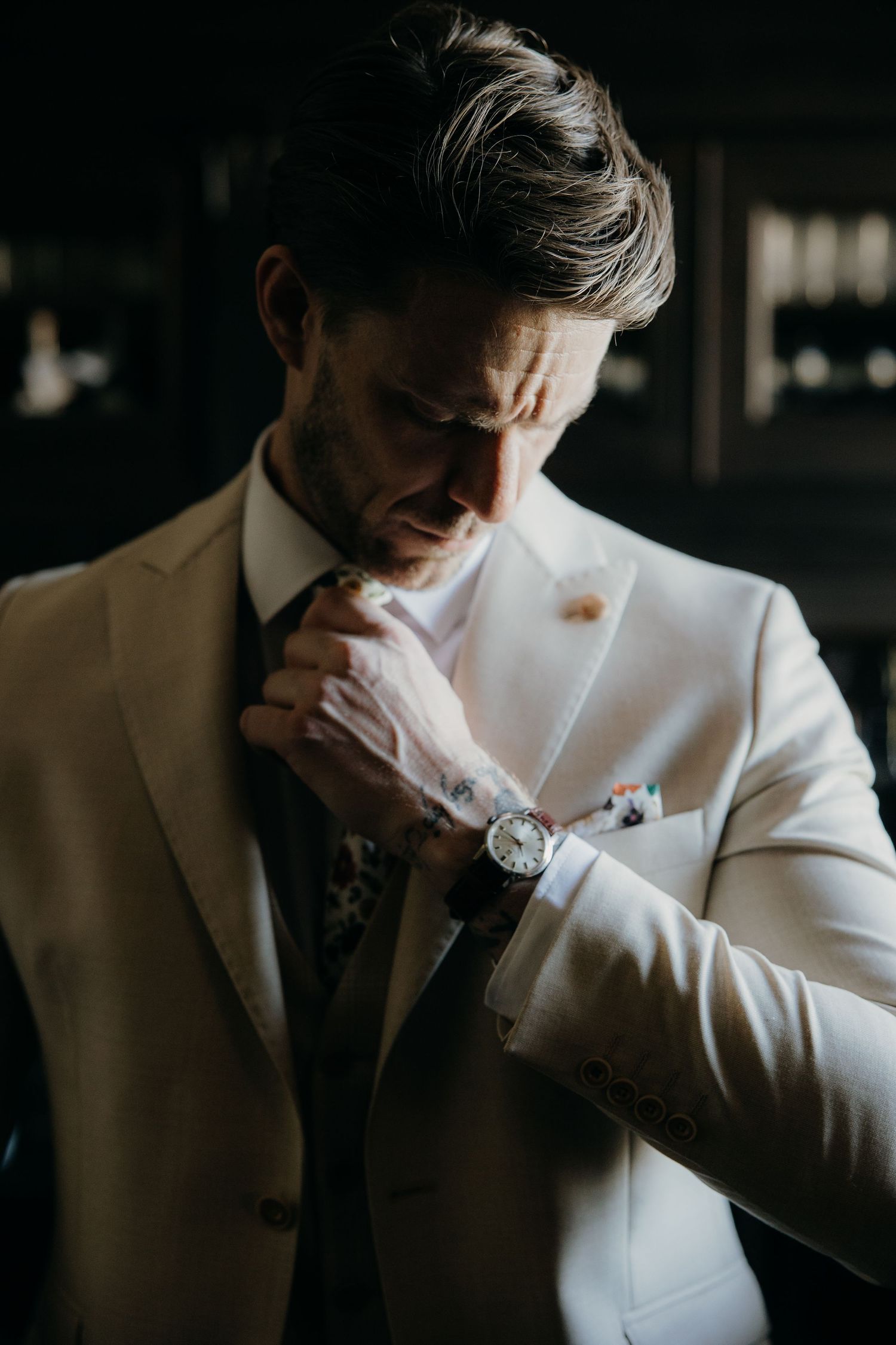 A well-dressed groom in a white suit adjusts his floral tie while wearing an elegant watch before his wedding ceremony.