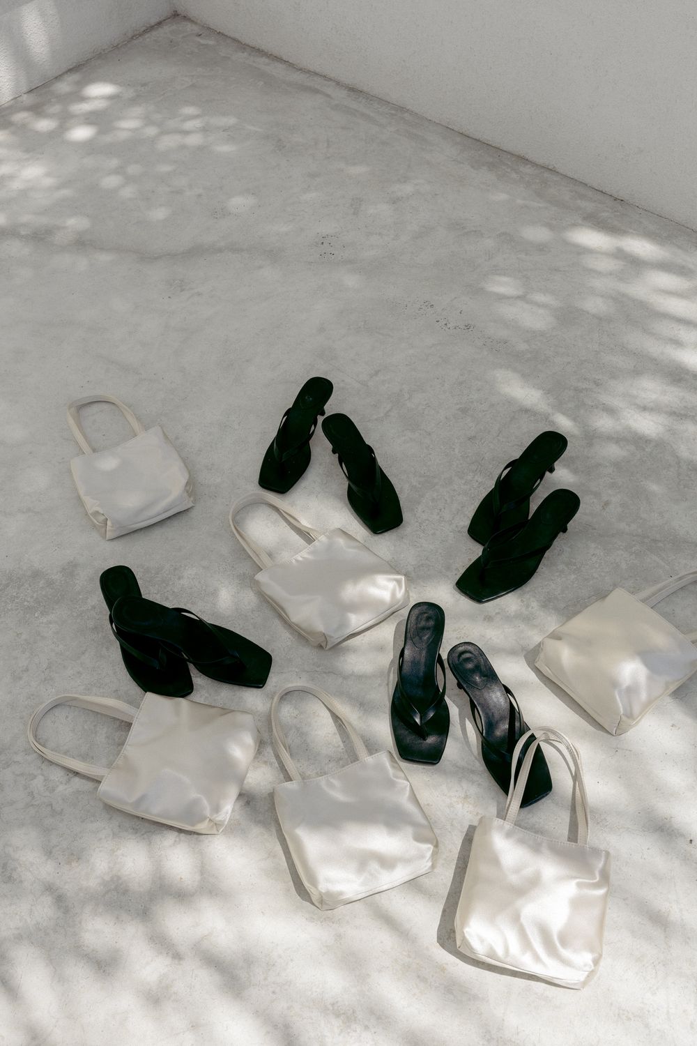 White tote bags and dark green sandals arranged on concrete floor with dappled light shadows.