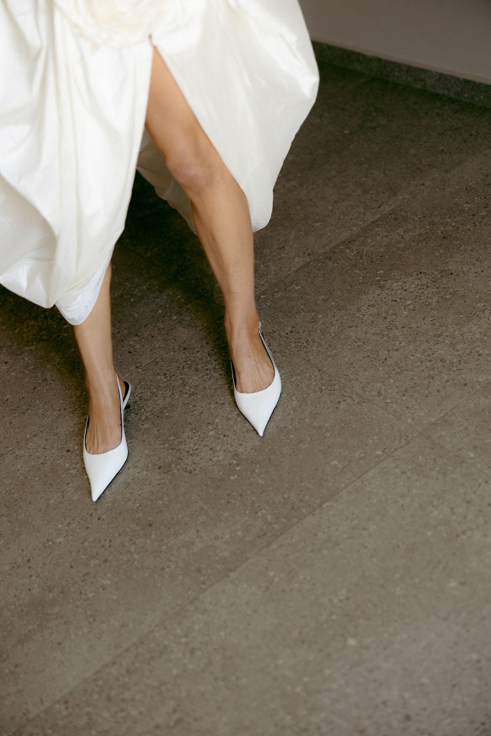 Close-up of bride's legs in pointed white mule heels beneath flowing satin wedding dress.