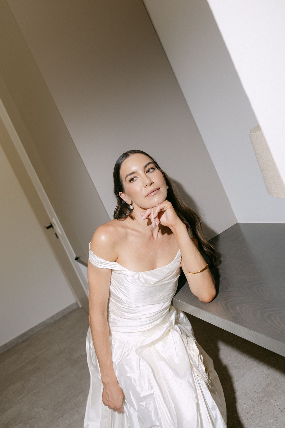 Elegant bride in an off-shoulder satin wedding dress sitting thoughtfully in soft natural light.