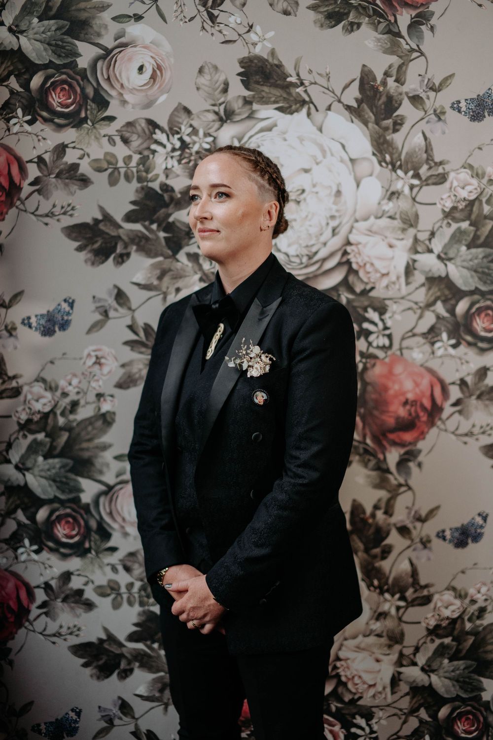 Person in black tuxedo with floral brooch stands before floral wallpaper.