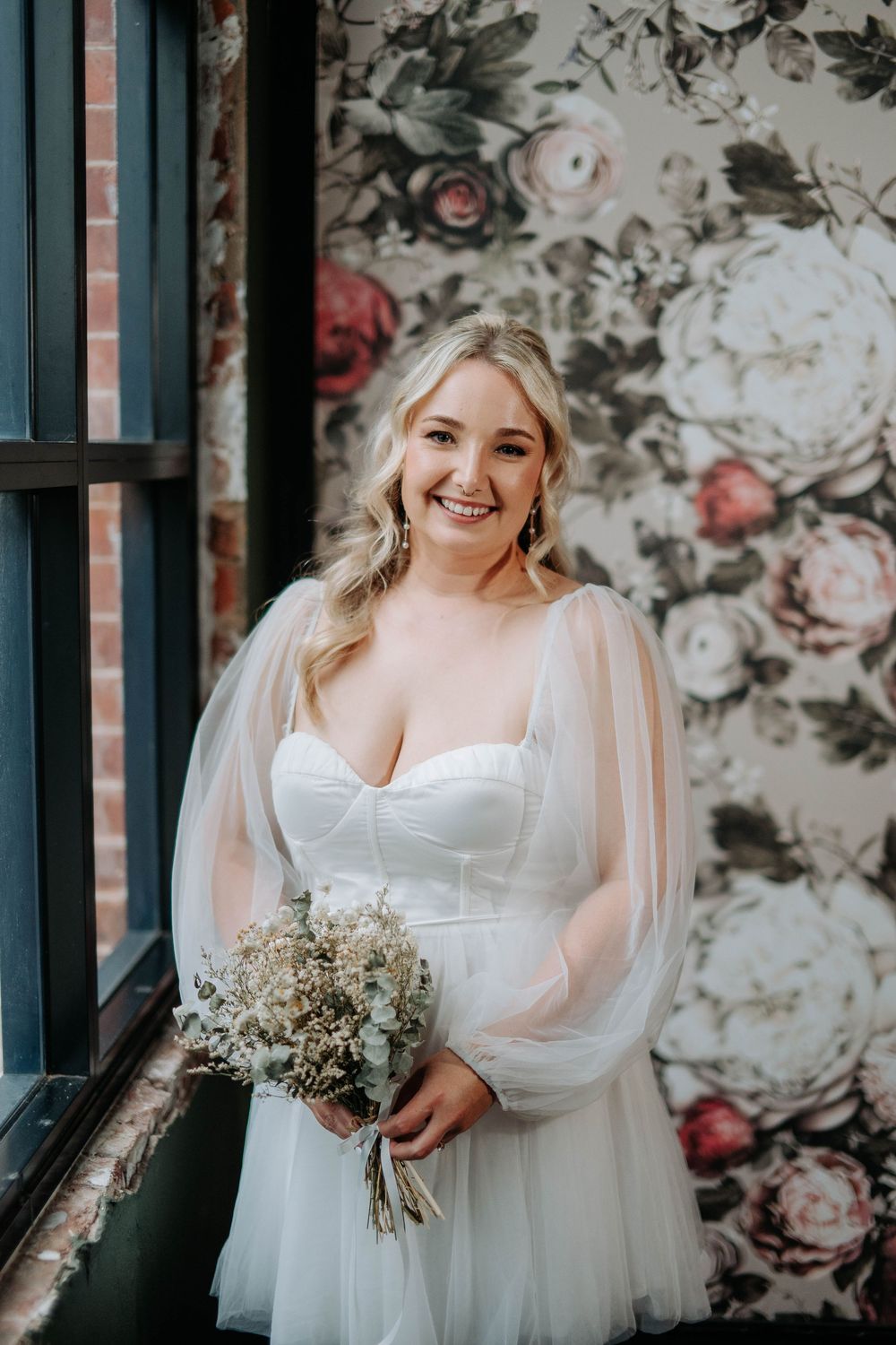 Smiling bride in white tulle gown holds bouquet before floral wallpaper.