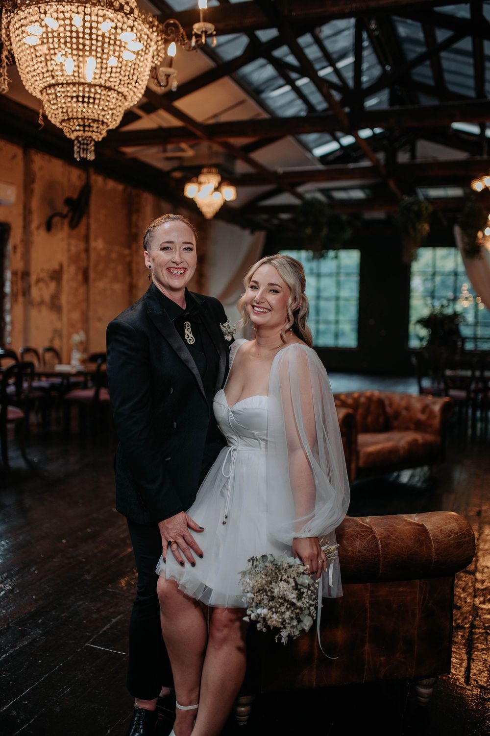 Two brides pose together in an elegant industrial wedding venue with crystal chandeliers and leather sofas.
