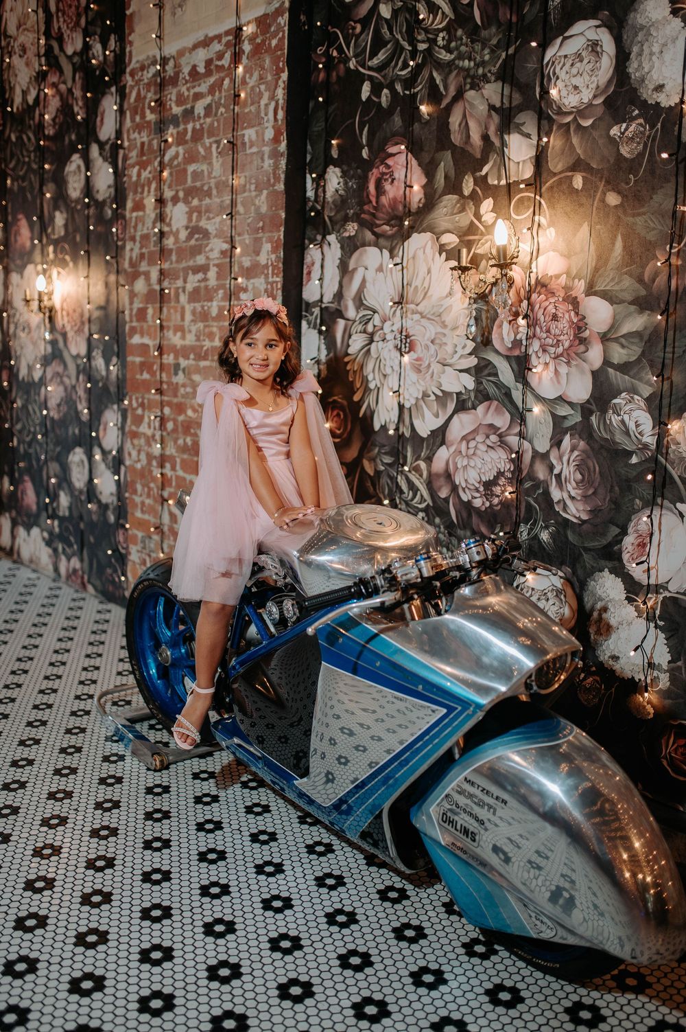 Young girl in pink tulle dress poses on a vintage blue motorcycle against floral wallpaper backdrop.