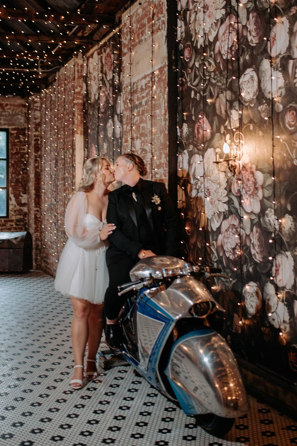 Bride and groom share a kiss beside a vintage motorcycle in a romantic venue with floral wallpaper and fairy lights.