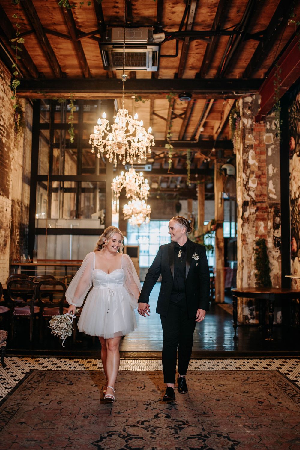 Two brides walk hand-in-hand through rustic venue with chandeliers, one in white mini dress, one in black suit.