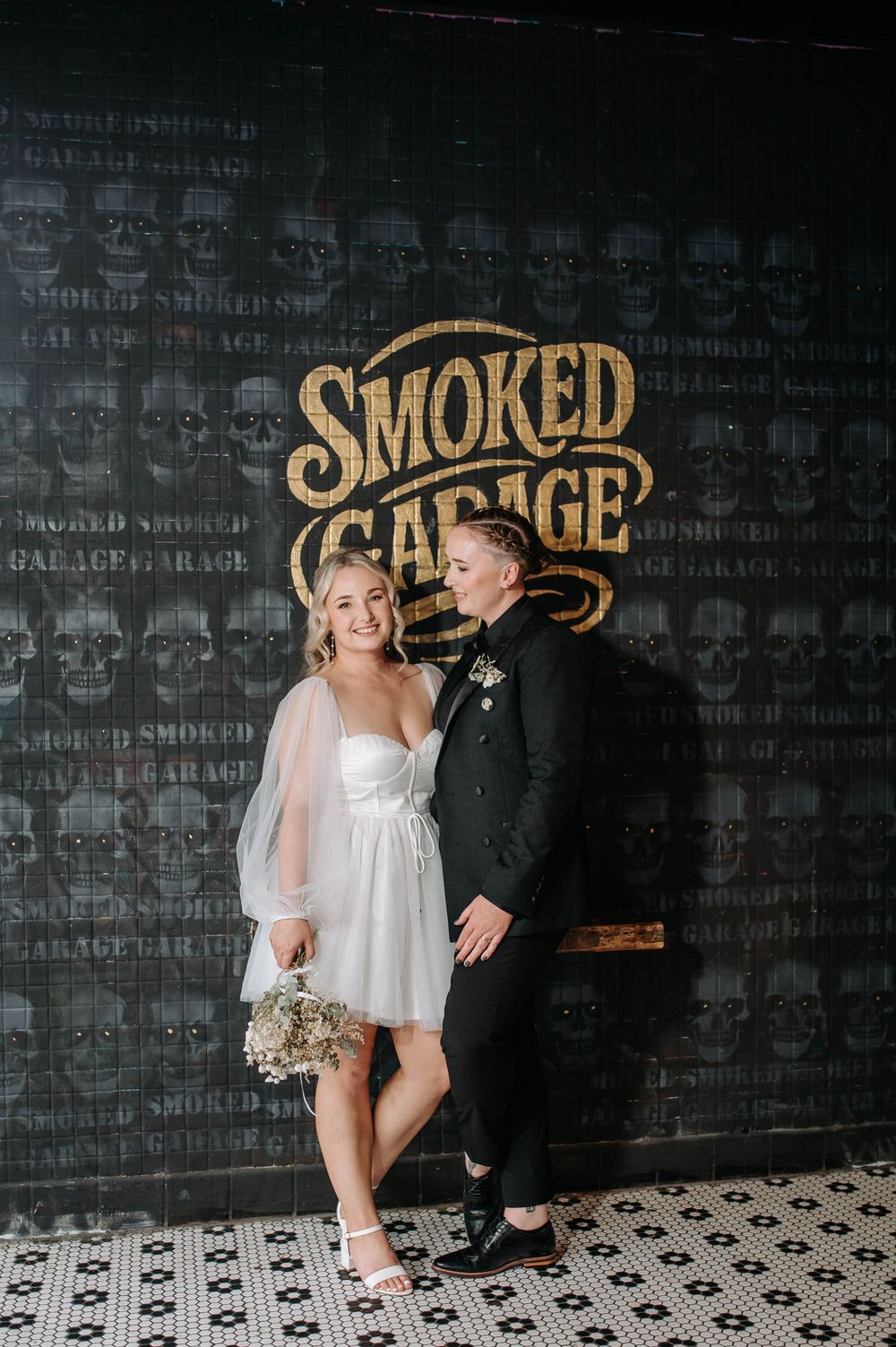 Two brides smile together in front of a dark Smoked Garage branded wall at their wedding reception.