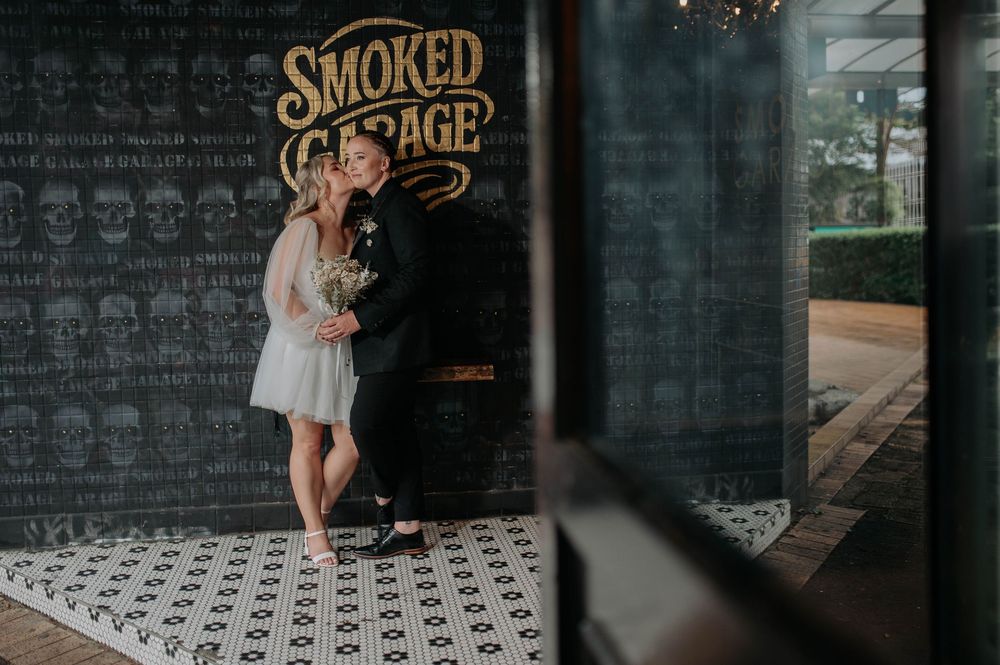Couple poses romantically in front of Smoked Garage sign on skull-patterned floor.
