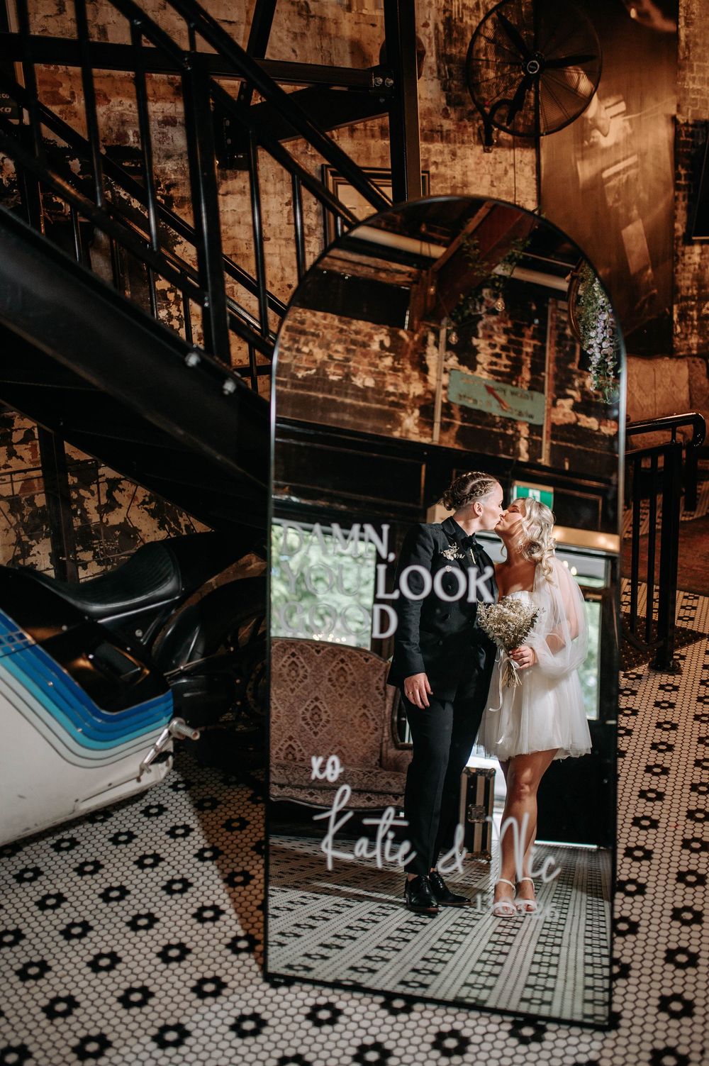 Bride and groom kissing reflected in a decorative mirror with script lettering in a rustic industrial wedding venue.