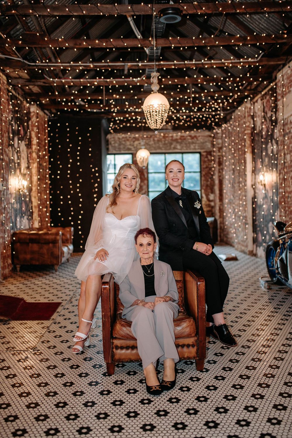 Two brides and elderly woman pose in fairy light-adorned wedding venue.