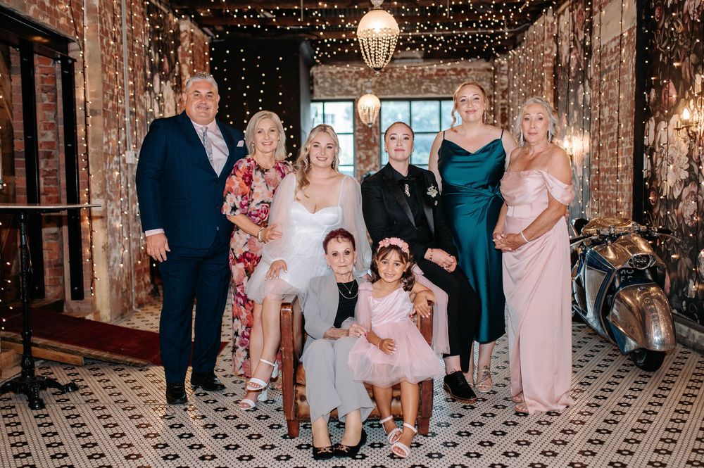 Wedding group photo with bride, groom, family, and flower girls posing in an elegantly decorated venue.