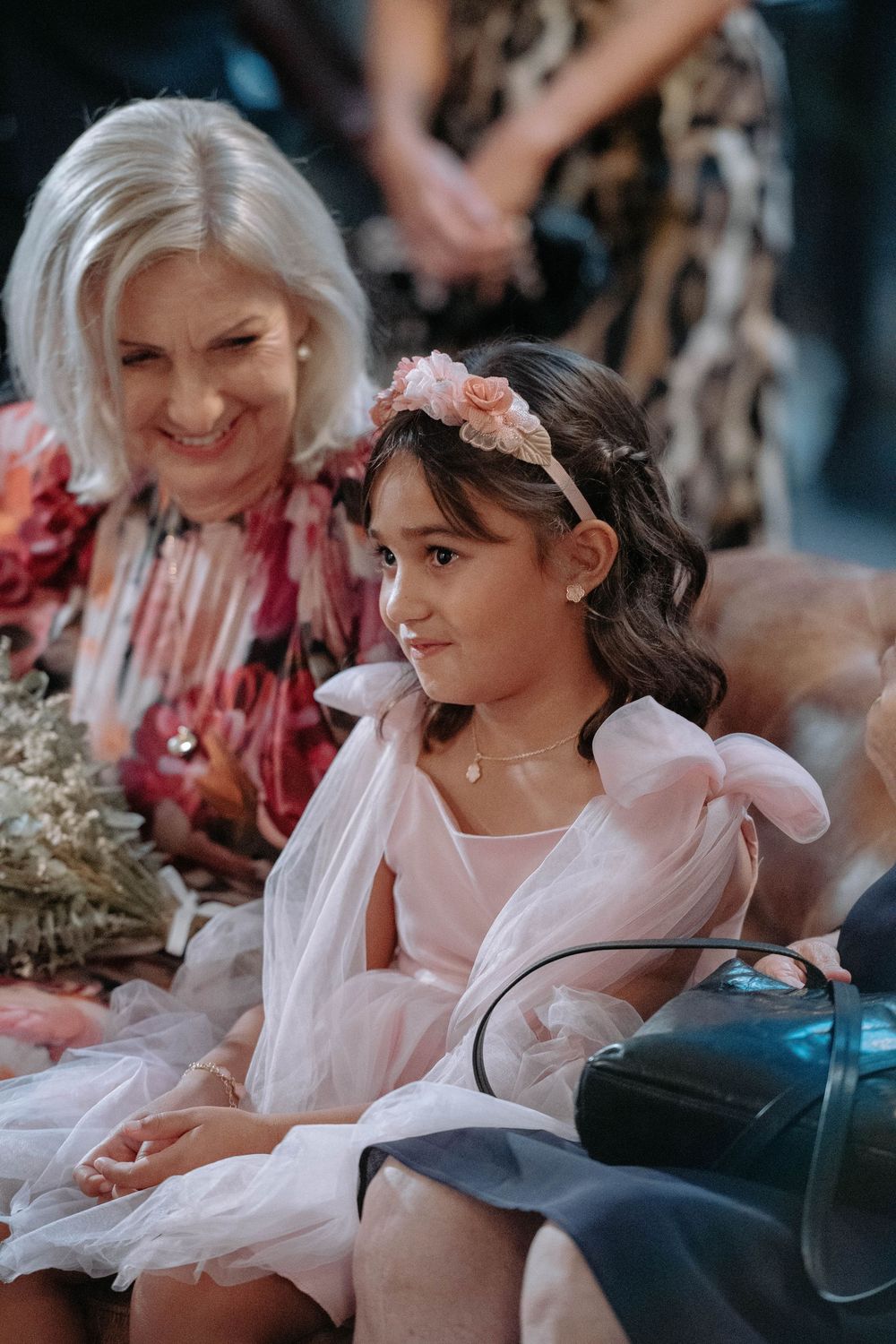 Young flower girl in pink dress with floral headband sits beside smiling elderly woman at wedding ceremony.