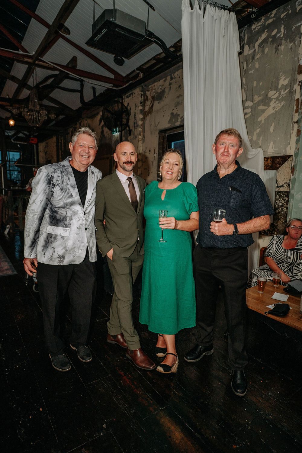 Four guests smile together at an indoor event, one woman in green dress holding champagne glass.