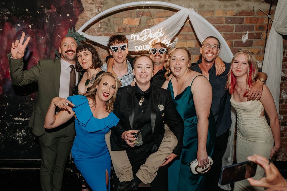 Group of wedding guests posing and celebrating at a reception with neon sign backdrop.