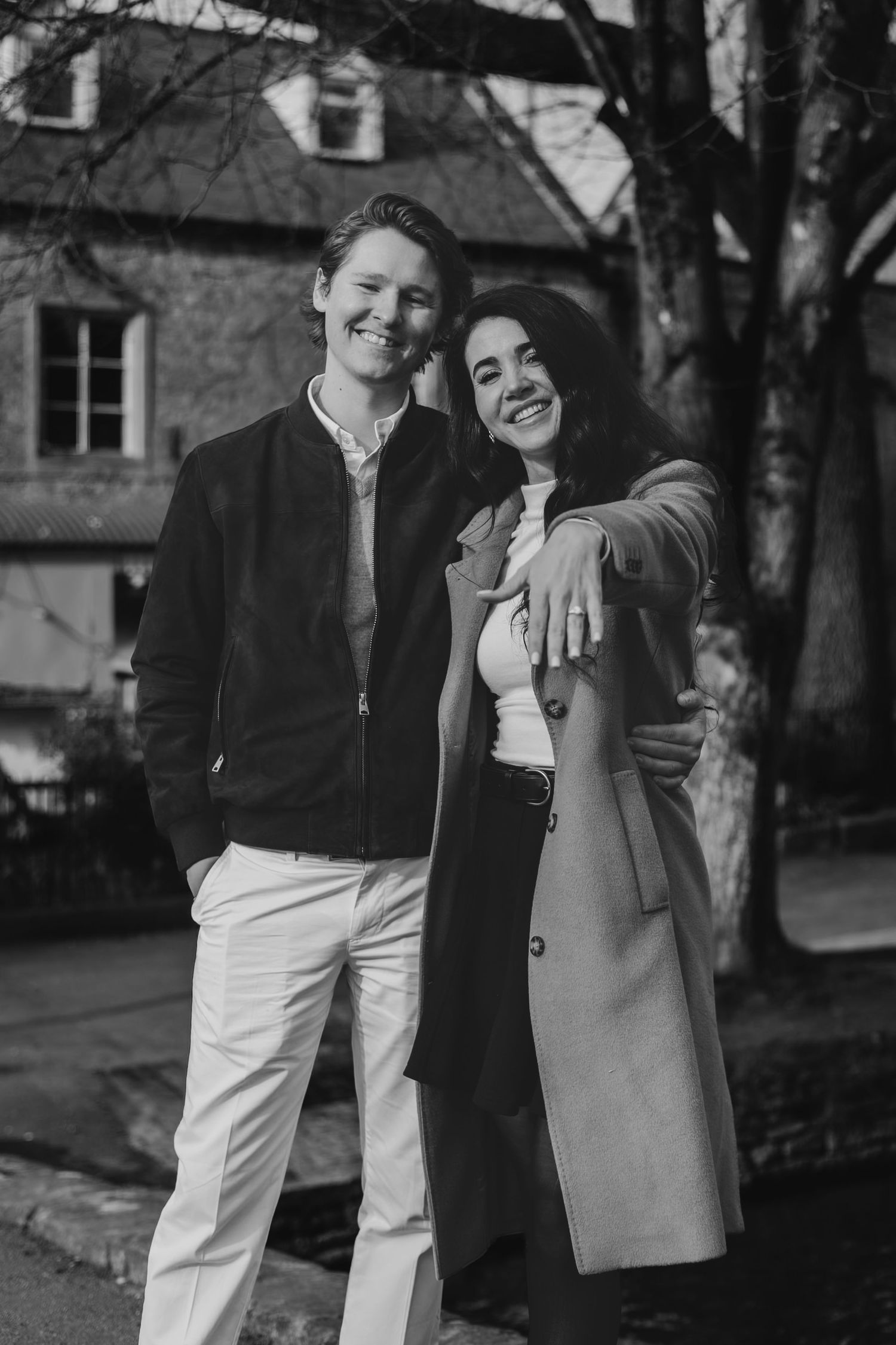 Smiling couple posing outdoors, woman showing engagement ring, historic building in background in bourton on the water.