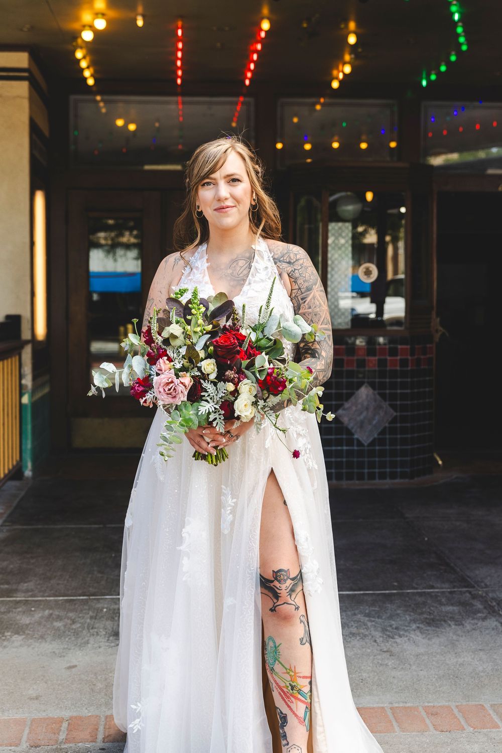 Tattooed bride in white lace gown with slit holds lush burgundy and blush bouquet outside a venue with colorful lights.