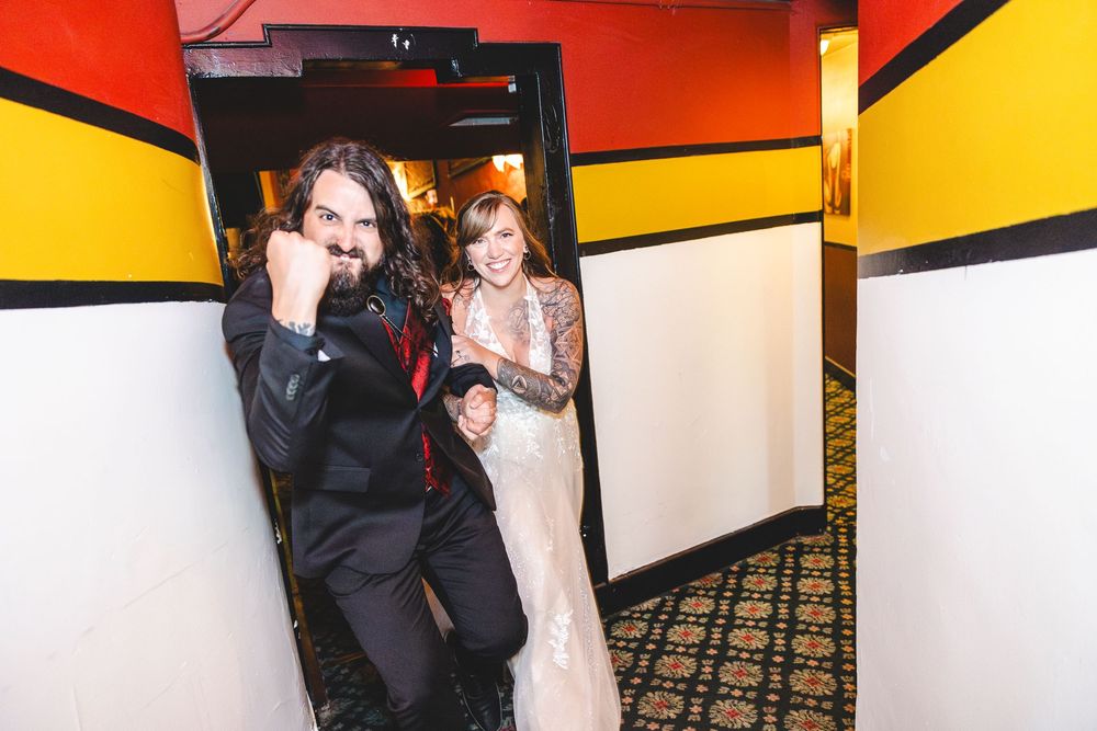 Excited newlyweds celebrating in a colorful hallway, groom in black suit and bride in lace wedding dress.