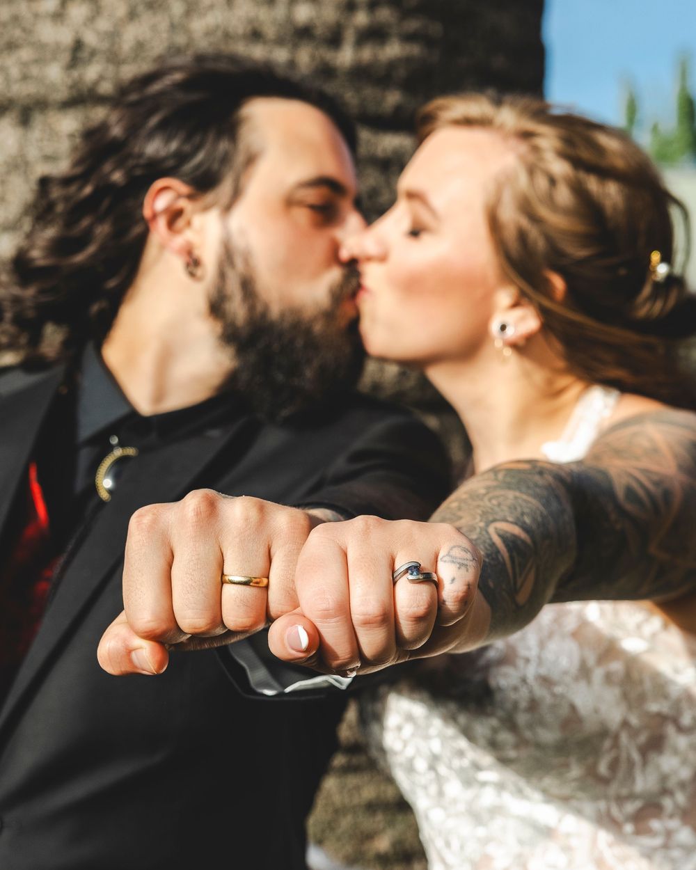 Newlyweds kissing while displaying wedding rings, bride with tattoos and groom with beard in outdoor portrait.