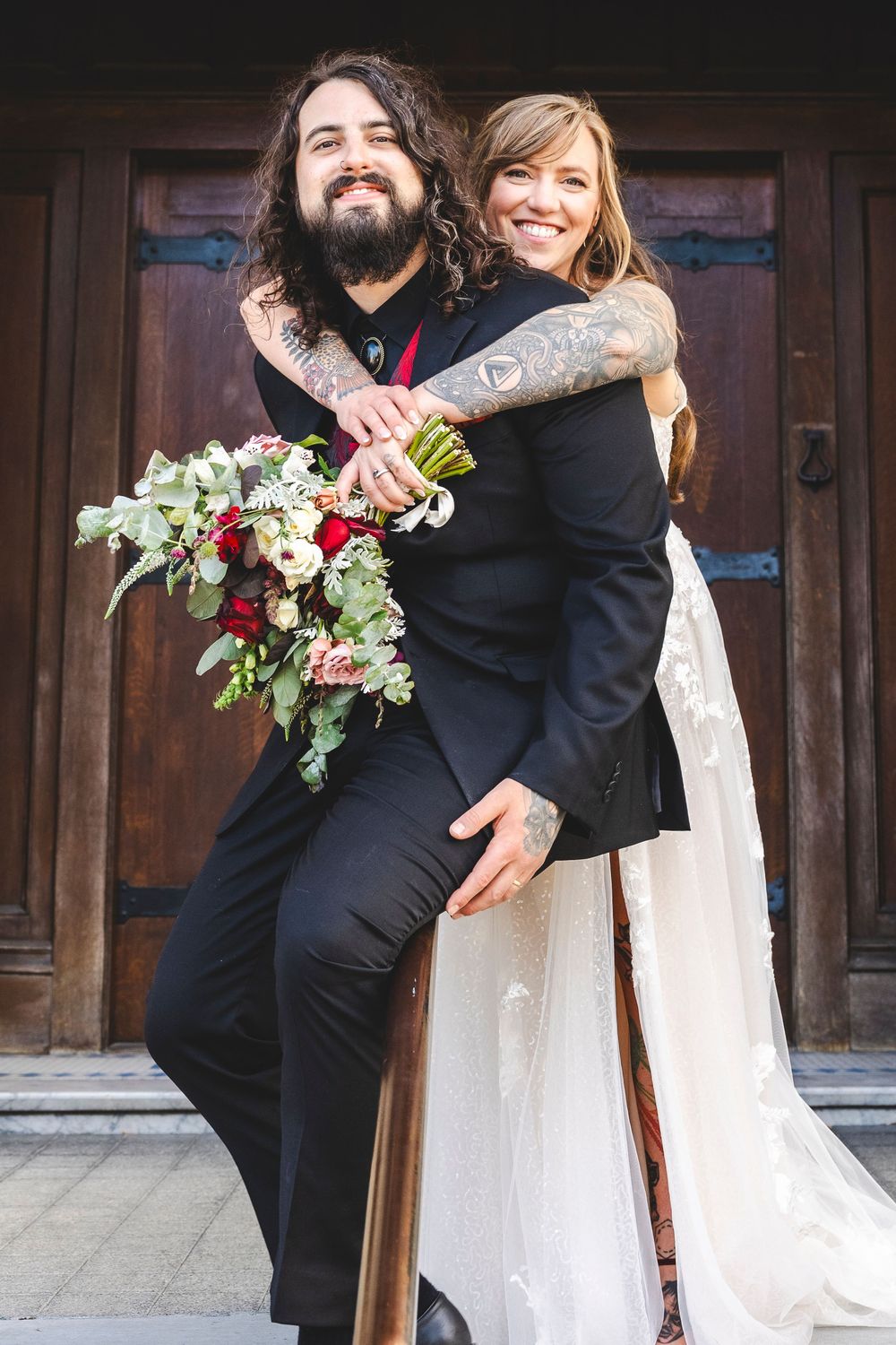 Tattooed groom in black suit holds bride in white dress with red bouquet outside wooden doors.