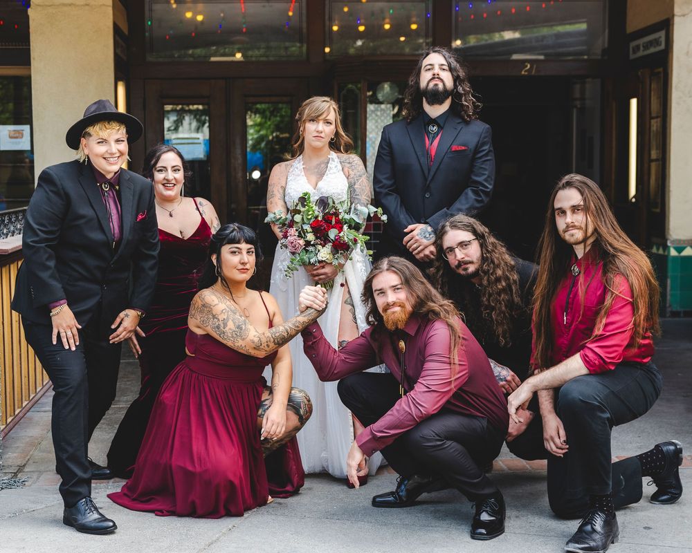 Wedding party in dark red and black attire poses outside venue with bride holding deep red bouquet.