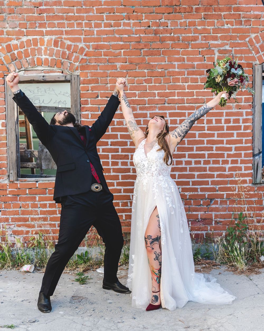 Tattooed bride and groom celebrate in front of brick wall, raising arms triumphantly holding bouquet on wedding day.