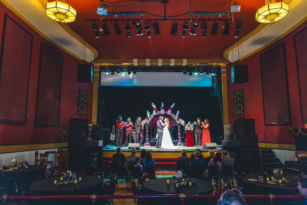 Wedding ceremony on a decorated theater stage with bridal party in red and white, guests seated in ornate venue.