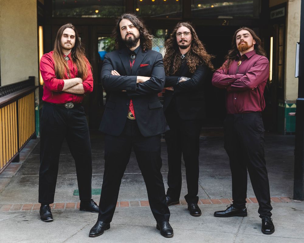 Four long-haired rock musicians pose confidently in dark formal attire outside a building.