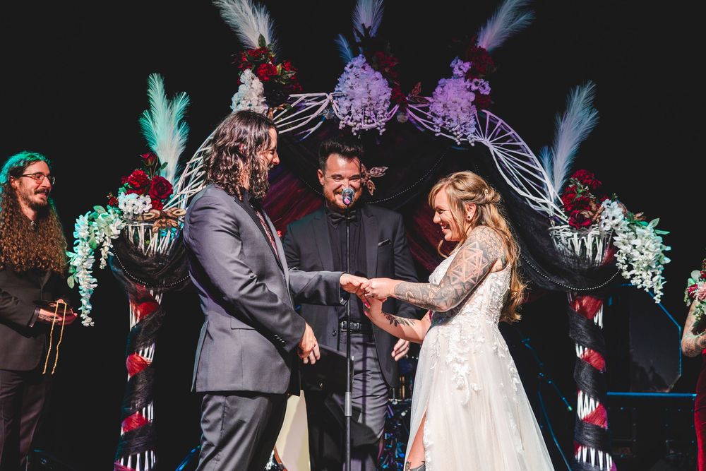 Couple exchanges rings at a colorful wedding ceremony with feathered arch and officiant on stage.