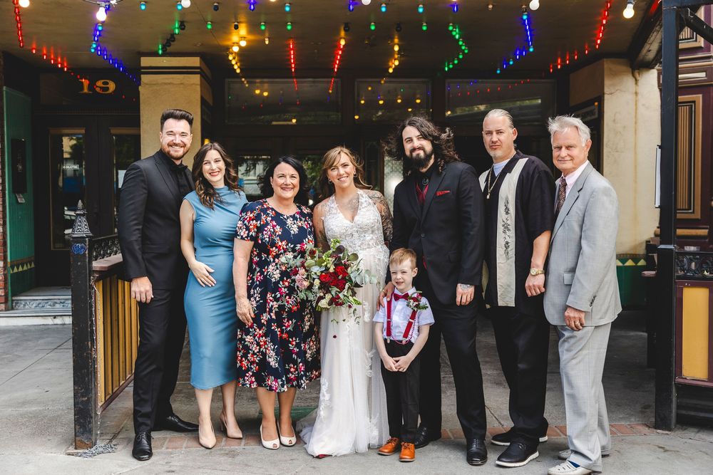 Wedding party group photo with bride in white gown, groom, and guests posing outside a venue with colorful string lights.