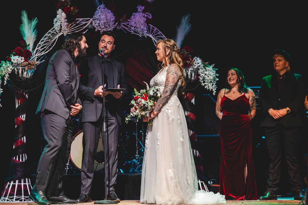 Couple exchanges vows at elegant wedding ceremony with officiant, bridesmaids in red velvet gowns, floral arch backdrop.