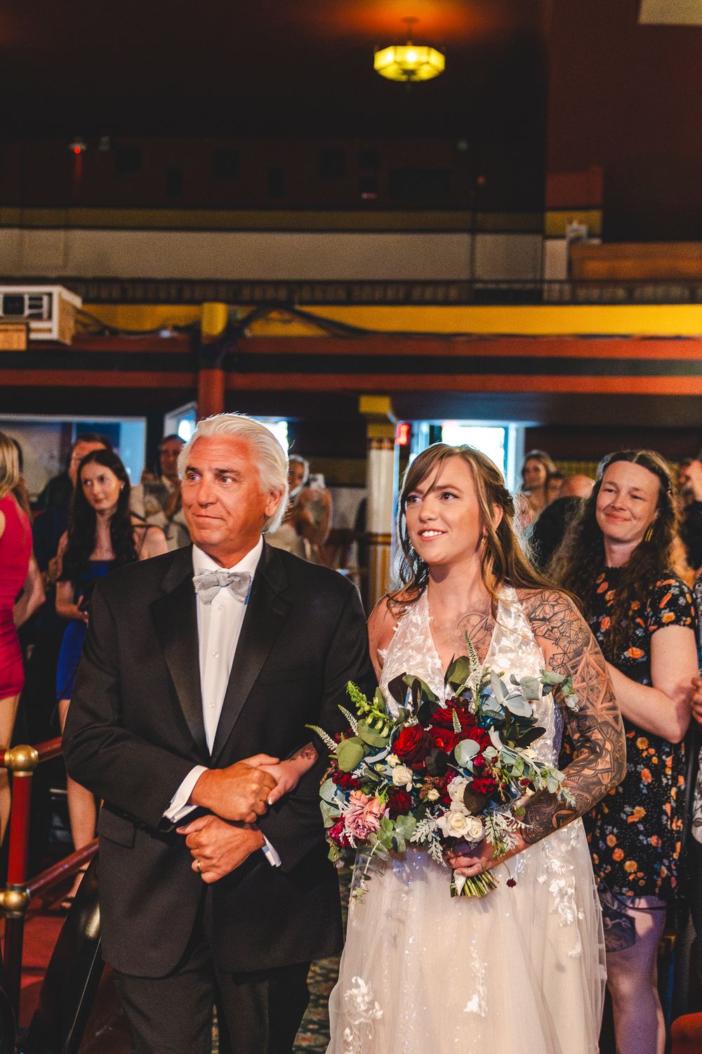 Bride in lace gown holding red bouquet walks down aisle with father in tuxedo at wedding ceremony.