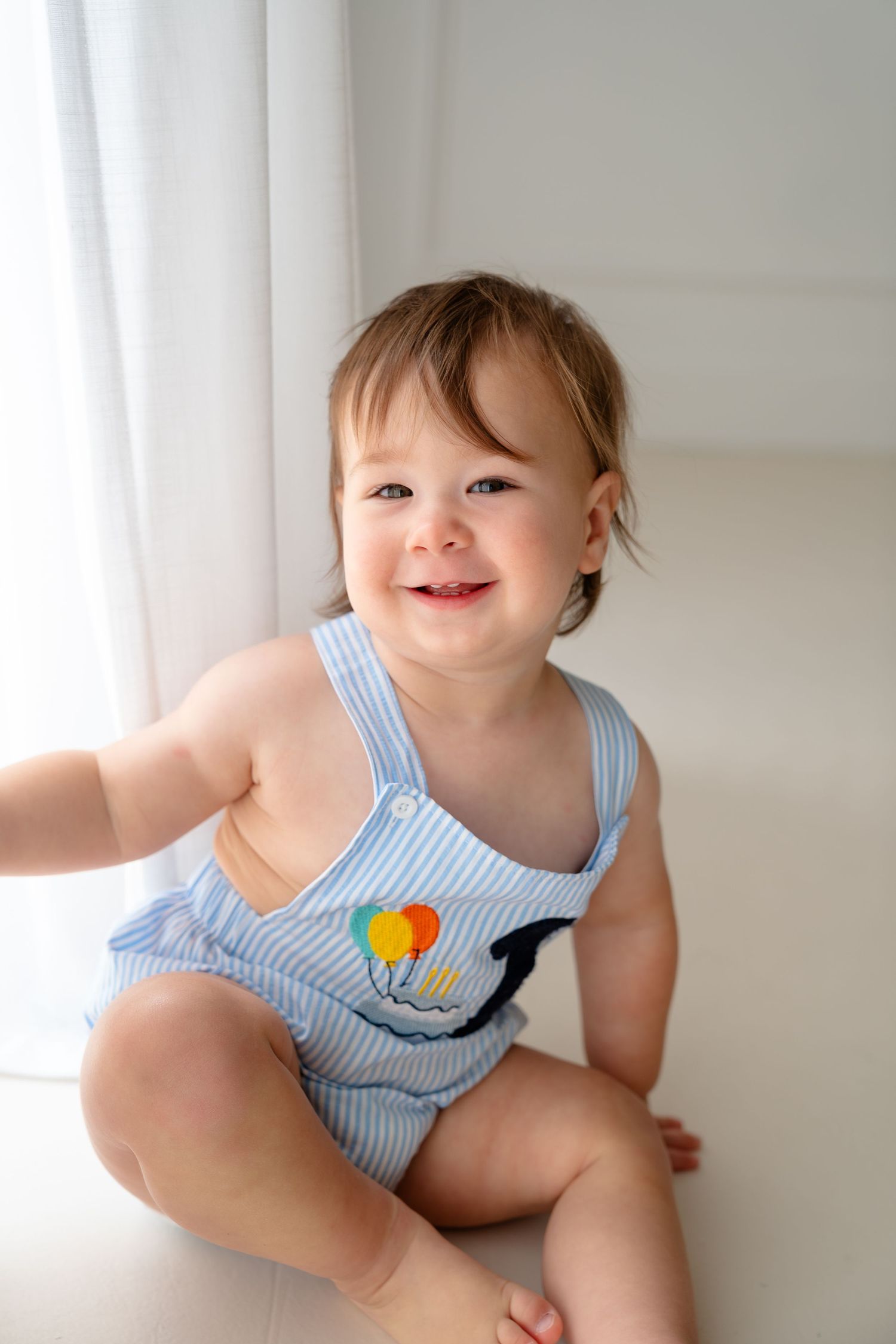 Smiling baby in blue striped romper with balloon applique sitting on white floor near curtains.