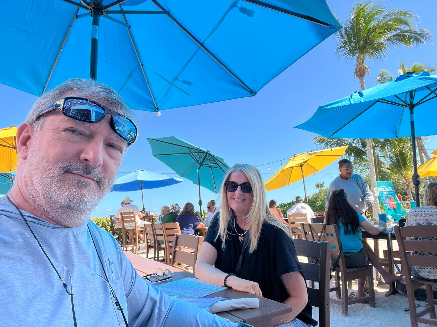 A couple smiles at an outdoor beachside restaurant with colorful blue and yellow umbrellas on a sunny day.