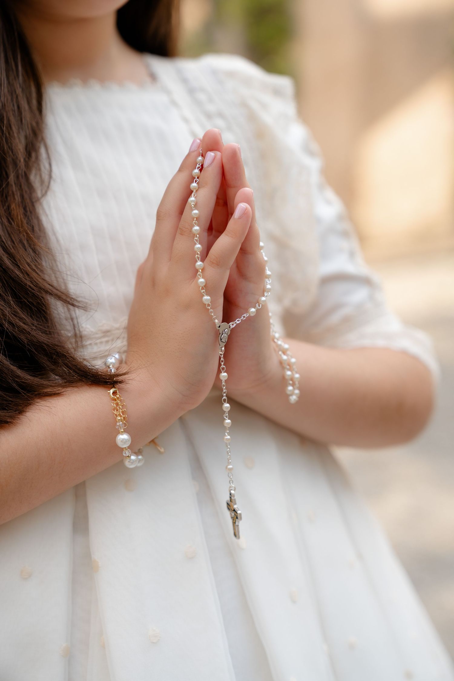 Young girl in white dress holds pearl rosary beads in prayer position for First Communion.